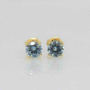 May include: A pair of gold stud earrings, each with a round, light blue gemstone. The gemstones are set in a classic four-prong setting. The earrings have a polished finish.