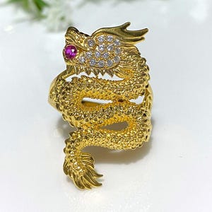 May include: A gold dragon ring with a red gemstone eye and clear rhinestones. The dragon is curled up with its tail wrapped around its body.