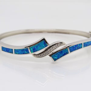 May include: A silver bangle bracelet with a blue opal design. The bracelet features a curved, wave-like pattern with multiple rectangular blue opal stones set in the silver.