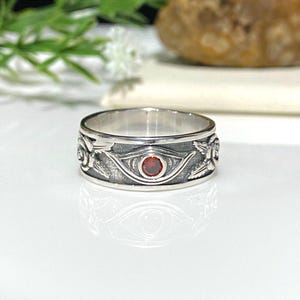 May include: A silver ring with an eye design and a red gemstone in the center. The ring is detailed with intricate carvings and has a polished finish.