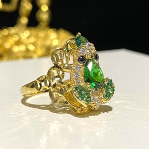 May include: A gold-colored ring featuring a detailed design with a large, pear-shaped green gemstone as the focal point. The ring is adorned with smaller green and white gemstones, and black accents. The ring's band has an openwork design.