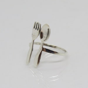 Puede incluir: Un anillo de plata con un dise&ntilde;o de tenedor y cuchara. El tenedor y la cuchara est&aacute;n unidos a la banda del anillo y se miran.