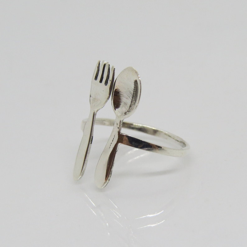 Puede incluir: Un anillo de plata con un dise&ntilde;o de tenedor y cuchara. El tenedor y la cuchara est&aacute;n unidos a la banda del anillo y se miran.