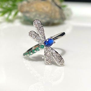 May include: A silver ring featuring a dragonfly design. The dragonfly has clear and blue gemstones on its wings and a green gemstone on its body.