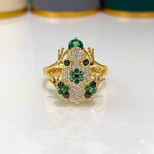 May include: A gold-toned ring shaped like a frog, featuring a body encrusted with clear white stones, two black eyes, and accents of green and black stones arranged in a floral pattern on the back and limbs, with a single green stone on the head.