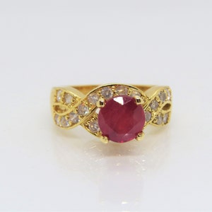 May include: A gold ring with a large red ruby gemstone surrounded by smaller clear gemstones. The ring has a twisted band design.