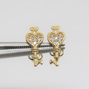 May include: A pair of gold key-shaped earrings with heart-shaped tops. Each heart is set with small, clear cubic zirconia stones.