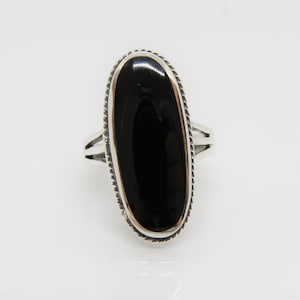 Peut inclure: Une bague en argent avec une pierre ovale noire sertie dans une bélière en argent. La bague présente un motif tressé délicat autour du bord de la bélière.