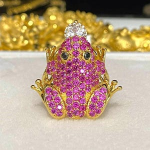 Peut inclure: Bague en forme de grenouille dorée, ornée d'une couronne et d'un corps recouvert de petites pierres roses. La grenouille a des yeux noirs et une grosse pierre transparente sur la tête. Fond flou.