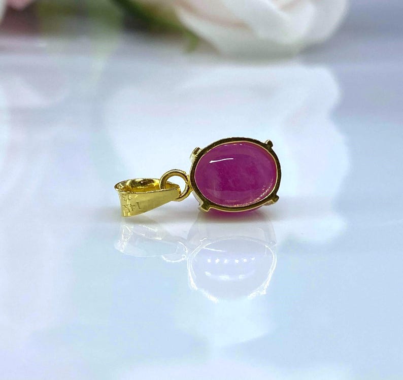 Puede incluir: Un colgante de oro con una piedra preciosa ovalada de color rosa. El colgante est&aacute; sobre una superficie blanca.