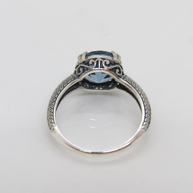 May include: A silver ring featuring a blue gemstone set within a filigree design. The band is textured with a repeating pattern.