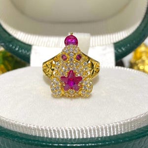 May include: A gold-toned ring featuring a whimsical design with a central star-shaped ruby and smaller ruby accents. The ring is encrusted with numerous small, clear gemstones, and a larger ruby tops the design. The ring is presented in a green and white jewelry box.