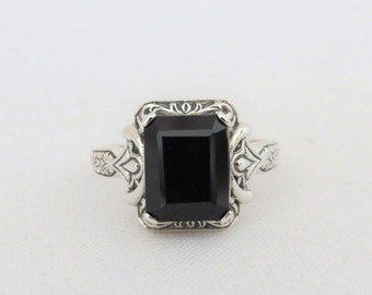 May include: A silver ring with an ornate design features a large, black, rectangular gemstone.