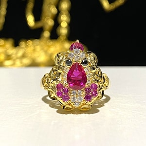 May include: A gold-colored ring featuring a central pear-shaped, deep pink gemstone. The ring is adorned with smaller pink and white gemstones, and black accents. The ring has an ornate, detailed design.