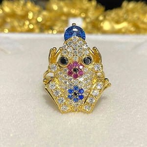 Peut inclure: Bague en forme de grenouille dorée, ornée d'une pierre bleue sur la tête. Le corps de la grenouille est incrusté de pierres précieuses claires et colorées, dont du noir, du rose et du bleu. La bague présente un design détaillé et orné.