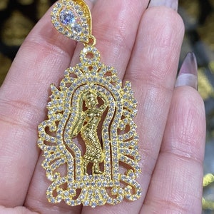 May include: Gold-toned pendant featuring a dancing figure within an ornate frame, encrusted with clear gemstones. The pendant has a bail with two clear gemstones. The design is intricate and detailed, with a polished finish.