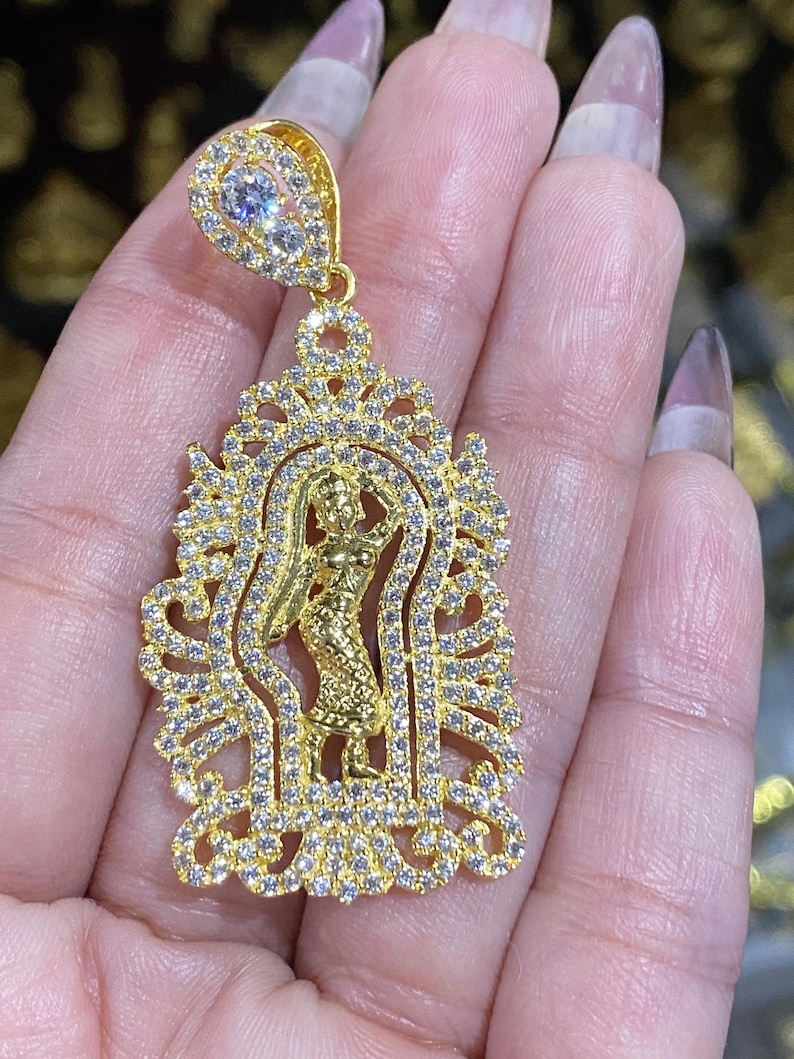 May include: Gold-toned pendant featuring a dancing figure within an ornate frame, encrusted with clear gemstones. The pendant has a bail with two clear gemstones. The design is intricate and detailed, with a polished finish.