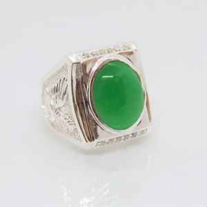 May include: A silver ring with a large, oval, bright green stone at the center. The ring has a rectangular face with small, clear stones around the green stone. The sides of the ring feature a detailed floral design.