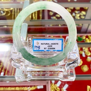 May include: A green jadeite jade bangle with a white background. The bangle is displayed on a clear plastic stand. The tag reads "NATURAL JADEITE JADE N: 230084".