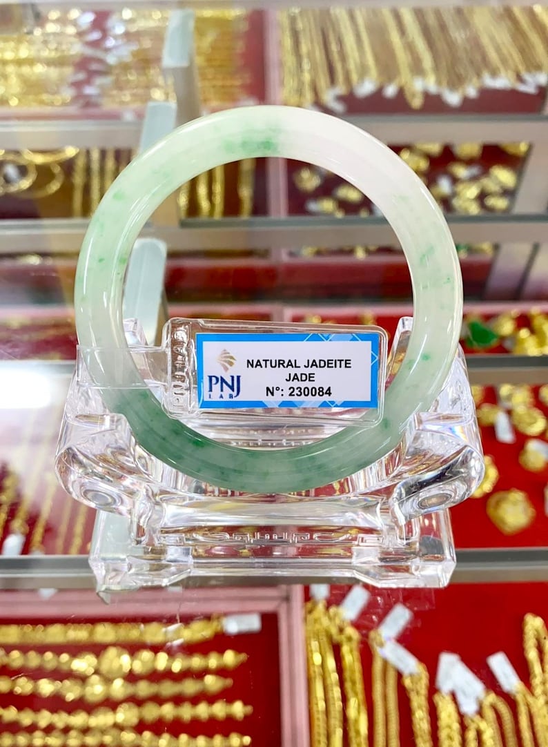 May include: A green jadeite jade bangle with a white background. The bangle is displayed on a clear plastic stand. The tag reads "NATURAL JADEITE JADE N: 230084".