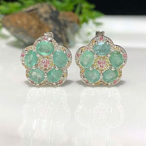 May include: A pair of flower-shaped earrings. Each earring features five oval-cut, light green emeralds surrounded by small pink and yellow gemstones. The earrings have a silver-colored metal setting and a post-back closure.