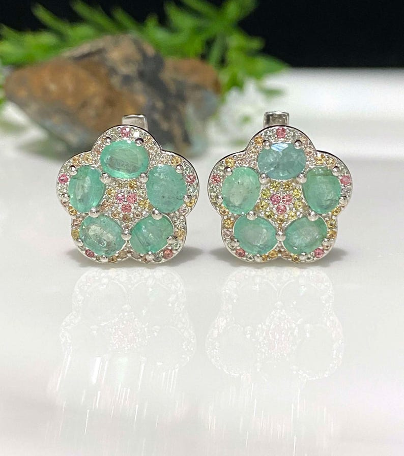 May include: A pair of flower-shaped earrings. Each earring features five oval-cut, light green emeralds surrounded by small pink and yellow gemstones. The earrings have a silver-colored metal setting and a post-back closure.