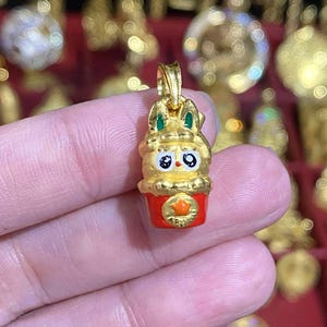 May include: A gold-colored pendant in the shape of a cartoon bunny with green accents on the ears. The bunny is atop a red cup with a gold star. The pendant is held in a person's hand.