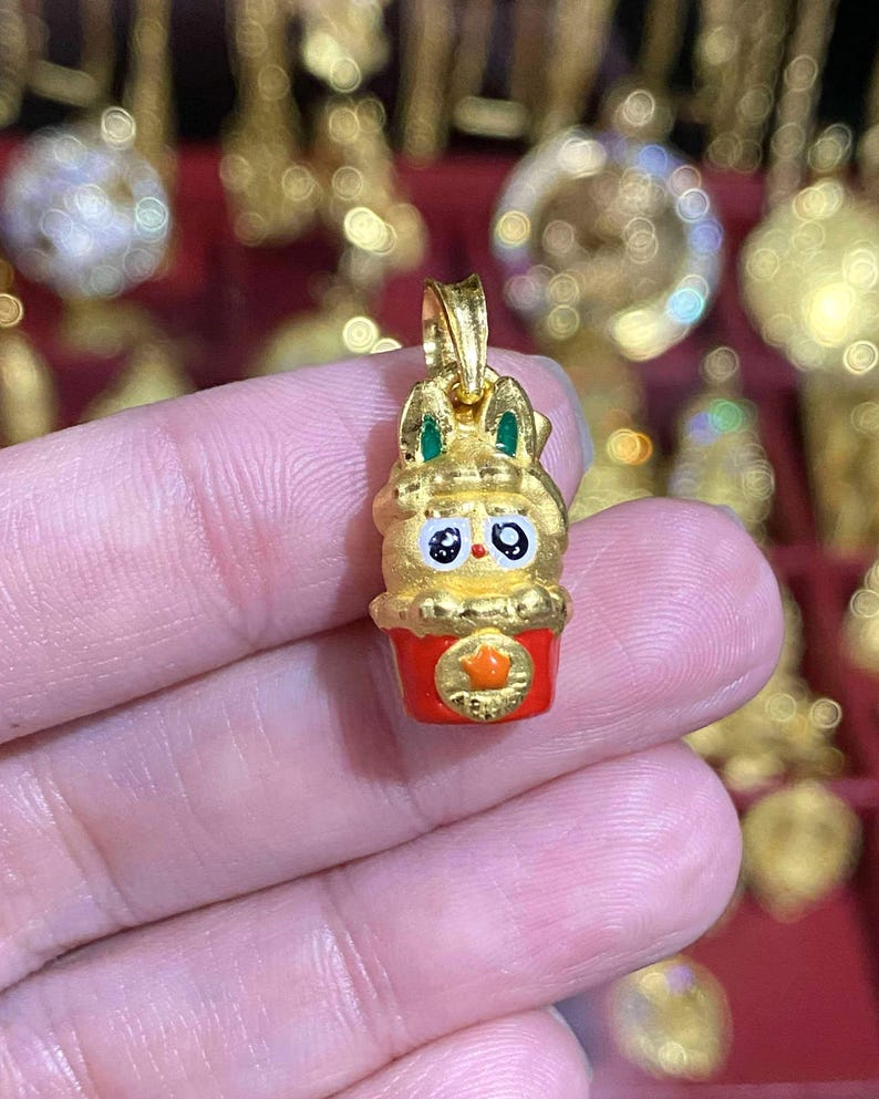 May include: A gold-colored pendant in the shape of a cartoon bunny with green accents on the ears. The bunny is atop a red cup with a gold star. The pendant is held in a person's hand.