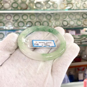 May include: A green jadeite jade bangle with a white background. The bangle has a label that reads "NATURAL JADEITE JADE N&deg;: 230084".