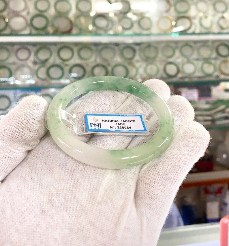 May include: A green jadeite jade bangle with a white background. The bangle has a label that reads "NATURAL JADEITE JADE N&deg;: 230084".