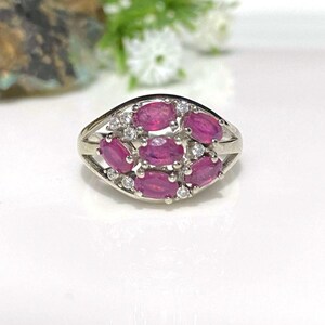May include: A silver ring featuring a cluster of oval-cut, deep pink gemstones, likely rubies, surrounded by smaller, clear gemstones. The ring has a split shank design and is set against a white background, showcasing the jewelry's details.