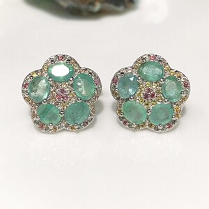 May include: A pair of flower-shaped stud earrings. Each earring features five oval-cut, light green gemstones surrounded by smaller pink and yellow stones. The earrings have a silver-colored metal setting.