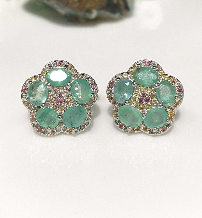 May include: A pair of flower-shaped stud earrings. Each earring features five oval-cut, light green gemstones surrounded by smaller pink and yellow stones. The earrings have a silver-colored metal setting.