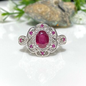 May include: A silver ring with a large oval ruby surrounded by smaller rubies and white diamonds. The ring has a delicate filigree design.