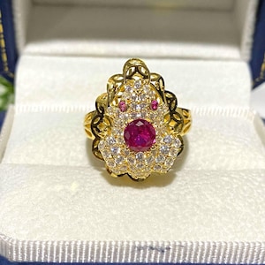 May include: A gold ring with a large red gemstone surrounded by smaller white gemstones. The ring has a delicate, floral design.