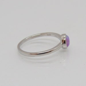 Puede incluir: Un anillo de plata con una piedra preciosa redonda de color p&uacute;rpura claro. El anillo tiene una banda delgada y un dise&ntilde;o simple y elegante. La piedra preciosa est&aacute; engastada en un bisel, sobre una superficie blanca.