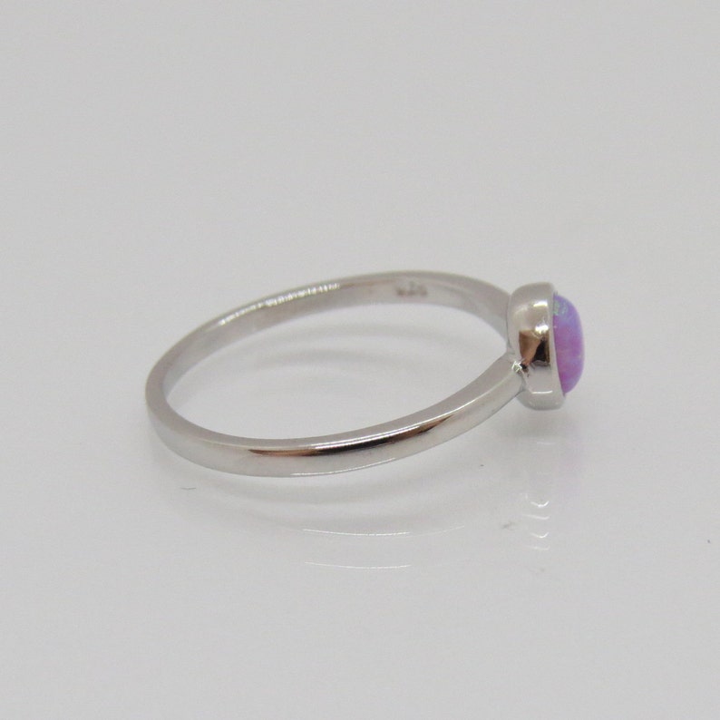 Puede incluir: Un anillo de plata con una piedra preciosa redonda de color p&uacute;rpura claro. El anillo tiene una banda delgada y un dise&ntilde;o simple y elegante. La piedra preciosa est&aacute; engastada en un bisel, sobre una superficie blanca.