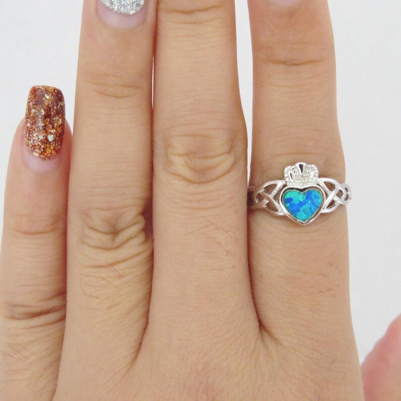 K&ouml;nnte beinhalten: Ein silberner Ring mit einem blauen Opal-Herz und einem keltischen Knotendesign. Der Ring wird an einem Finger getragen.