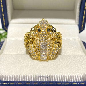 May include: A gold ring with a detailed design featuring a large, clear stone in the center and smaller, clear stones surrounding it. The ring is set in a gold band.