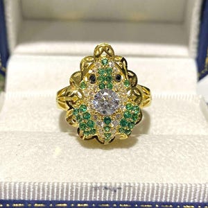 Puede incluir: Un anillo dorado con un diseño de rana. La cabeza de la rana está adornada con una gran piedra preciosa transparente rodeada de piedras blancas y verdes más pequeñas. El anillo se presenta en una caja azul y blanca.