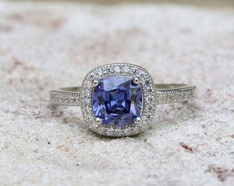 Op de afbeelding: Een zilveren ring met een grote, vierkante, blauwe edelsteen omringd door een halo van kleinere, witte edelstenen.