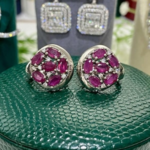 May include: A pair of silver-toned rings, each featuring a cluster of oval-cut, deep red gemstones and small, clear accent stones. The rings are displayed on a textured, dark green surface, with a pair of square, diamond-like earrings in the background.