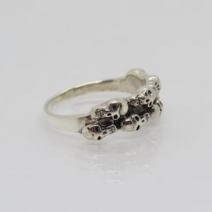 May include: Silver ring with a band of skulls. The skulls are detailed and have a slightly gothic aesthetic.
