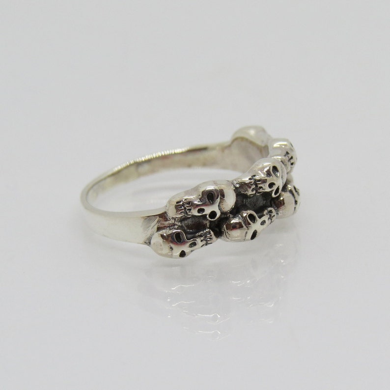 May include: Silver ring with a band of skulls. The skulls are detailed and have a slightly gothic aesthetic.