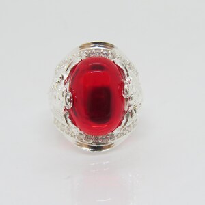 May include: A silver ring with a large, red, oval-shaped gemstone set in a decorative, silver setting with small, clear stones around the edge.