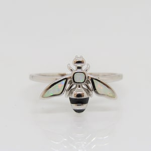 Può includere: Un anello d'argento con un design a forma di ape. Le ali dell'ape sono in opale bianco e il suo corpo è bianco e nero.