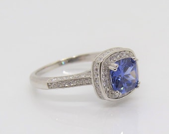 Op de afbeelding: Een zilveren ring met een grote, vierkante blauwe edelsteen omringd door kleinere, heldere edelstenen.