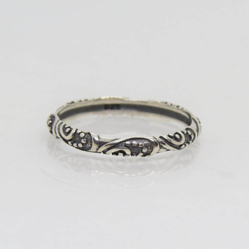 May include: A silver ring with a delicate, intricate design featuring swirls and dots. The ring is thin and has a polished finish.