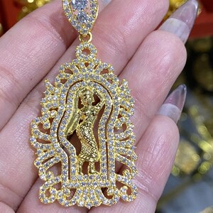 May include: A gold-colored pendant featuring a detailed design of a figure within an ornate frame, encrusted with small, clear gemstones. The pendant has a teardrop-shaped top with two larger gemstones. The jewelry is held in a hand.