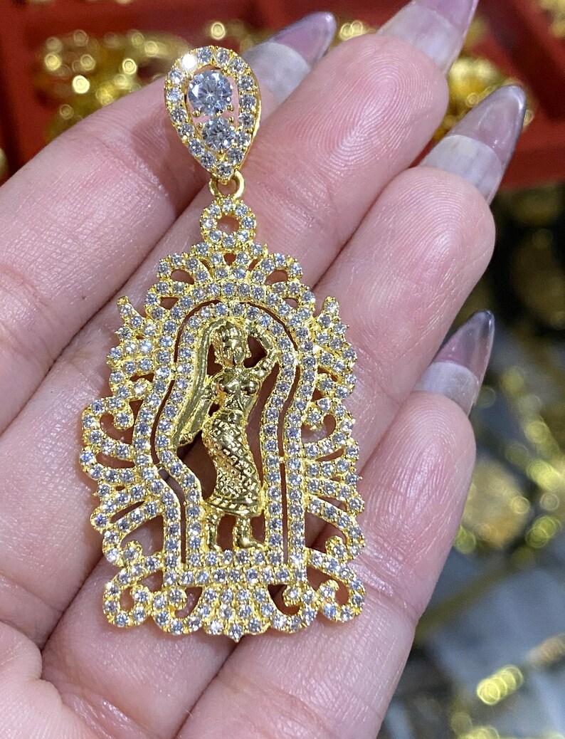 May include: A gold-colored pendant featuring a detailed design of a figure within an ornate frame, encrusted with small, clear gemstones. The pendant has a teardrop-shaped top with two larger gemstones. The jewelry is held in a hand.